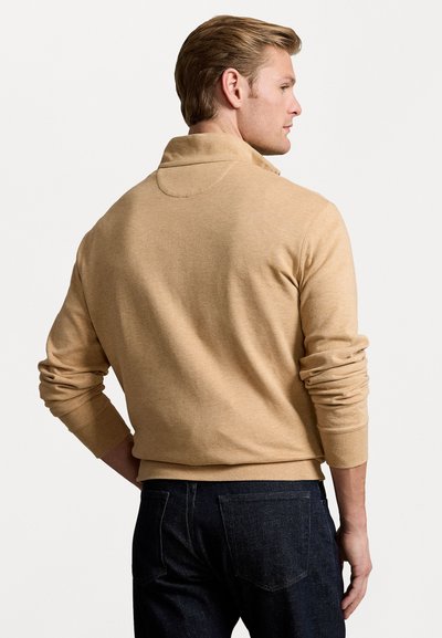Beige stickad tröja med krage, långärmad och med ribbade ärmmuddar, i kombination med mörka jeans. Slät textur och enkelt design.