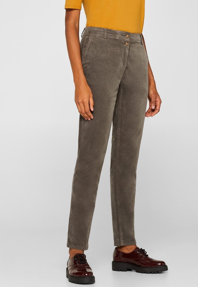 Personne portant un pantalon en velours côtelé vert olive, un haut à manches courtes moutarde et des chaussures à lacets vernies rouge foncé, debout devant un fond uni.