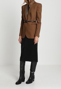 Blazer marrone su misura con una cintura nera, abbinato a una gonna nera in satin e stivali al ginocchio in pelle nera. Design semplice e strutturato.