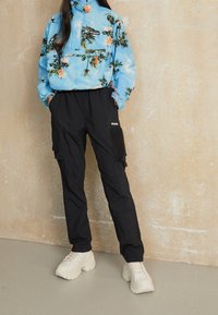 Person som bär en blå blommig Columbia-jacka, svarta cargo-byxor och vita klumpiga sneakers, står mot en beige vägg.