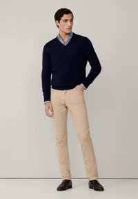 Pullover de V da marinha, camisa colarinho clara azul por baixo, calças slim-fit bege, e sapatos loafers escuros. Textura suave com design simples.