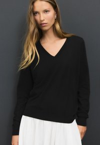 Maglione nero a scollo a V con maniche lunghe, texture morbida e polsini a coste, abbinato a una gonna bianca. Colori solidi senza motivi.