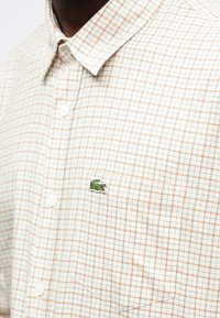 Srajca s potiskom karirastega vzorca v beli in svetlo rjavi barvi, z zelenim vezenim logotipom krokodila na prsnem žepu. Iz bombažnega materiala.