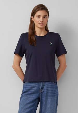 Vrouw draagt een marineblauw T-shirt met korte mouwen en een kleine witte bloemenborduursel op de borst, samen met een blauwe spijkerbroek, staand tegen een effen muur.