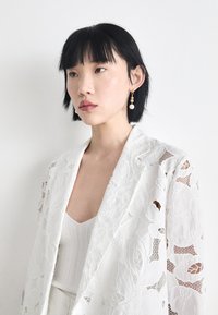 White embroidered lace jacket with floral patterns, worn over a light gray ribbed top. Features delicate earrings with pearls and gold accents.