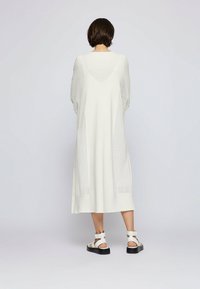 Abito lungo e oversize di colore bianco con dettagli in maglia testurizzata sulle maniche e sui lati. Abbinato a sandali bianchi con suola nera.