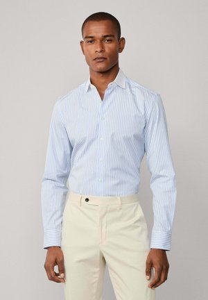 Mann in einem hellblauen und weißen gestreiften Hemd und cremefarbener Hose vor einfarbigem Hintergrund.