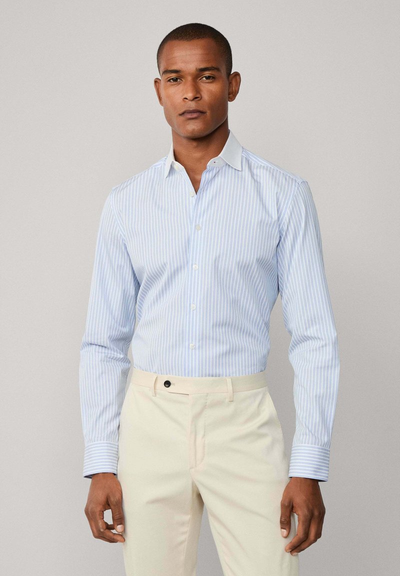 Uomo che indossa una camicia a righe verticali leggera blu e bianca e pantaloni color crema su uno sfondo neutro.