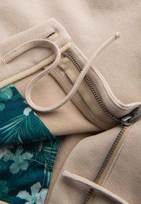 Textil beige con un acabado suave, que presenta un interior con un patrón floral. Incluye un cierre con cremallera y un detalle de cordón decorativo.
