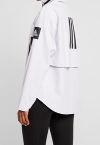Persona vestida con una chaqueta blanca de Adidas con parches de logo negro en la manga y la parte trasera, combinada con leggings negros, de pie de lado.