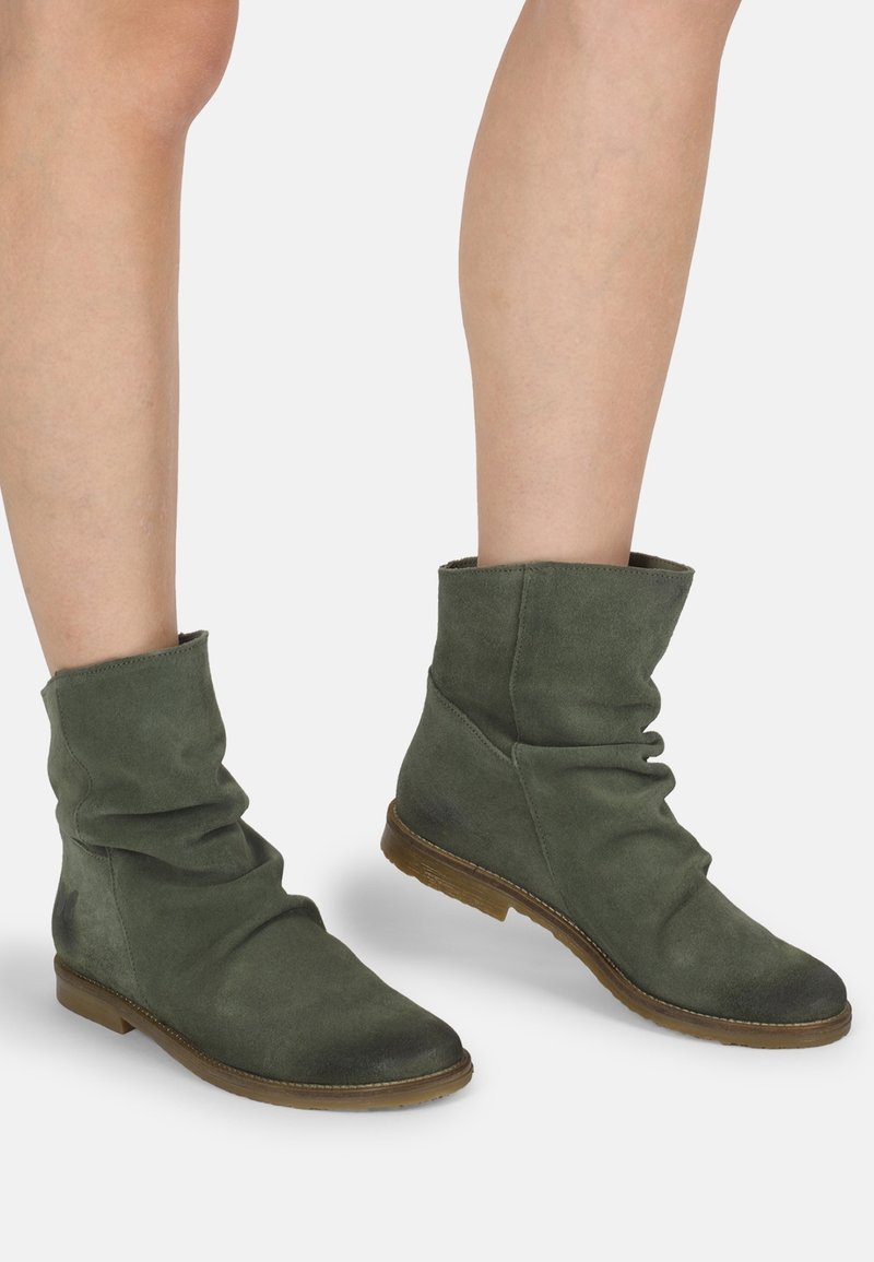 Bottes décontractées mi-mollet en daim vert foncé avec semelles plates marron portées sur des jambes nues sur fond blanc.