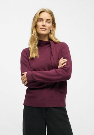 Burgundowy sweter z wysokim kołnierzem, długimi rękawami i prążkowanymi mankietami. Wykonany z miękkiego materiału, z boku z wiązaniem i luźnym krojem.