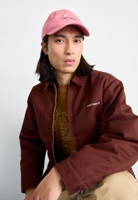 Brun zip-up jacka och brun grafisk t-shirt under, kombinerat med en rosa keps med grön text. Släta texturer och avslappnad passform.