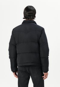 Chaqueta de plumas negra con acolchado horizontal, cuello puntiagudo y tela texturizada, llevada sobre jeans oscuros. Vista desde la parte posterior.