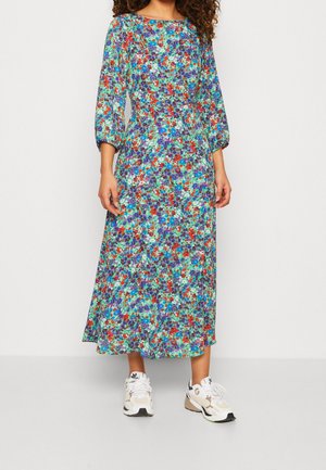 Femme portant une robe fleurie à manches longues et longueur cheville dans des tons de bleu, rouge et violet, associée à des baskets blanches.