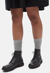 Bottes chevilles en cuir noir avec lacets et fermeture éclair latérale. Associées à des chaussettes côtelées grises. Texture lisse, bout arrondi et semelle plate.
