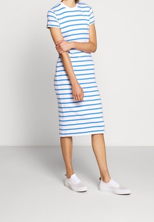 Personne portant une robe midi blanche avec des rayures horizontales bleues et des baskets blanches, debout devant un fond clair uni.