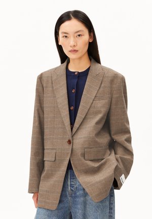 Vrouw draagt een bruin geruite blazer over een navy blauw gebuttonde trui en blauwe jeans, staand met één hand in haar zak tegen een witte achtergrond.