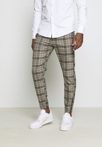 Man som bär gröna och beige rutiga byxor, vit skjorta med knappar och vita Adidas-sneakers står framför en enfärgad bakgrund.