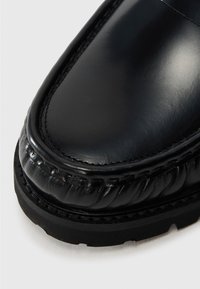 Zapato de cuero negro con un acabado brillante, punta redonda y detalle texturizado de costura a lo largo del borde. Diseñado con una suela de goma negra duradera.