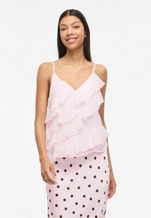 Hosszú, sötét hajú nő világos rózsaszín fodros ujjatlan felsőben és világos rózsaszín, sötét pöttyös szoknyában, mosolyogva, állva.