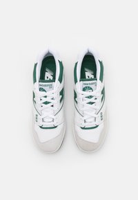 Sneakers bianchi con accenti verdi, materiali in pelle e camoscio, lacci prominenti e branding "New Balance 550" sulla linguetta e sul lato.