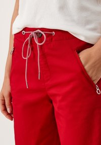 Pantalones rojos con cintura de cordón, cuentan con bolsillos con cremallera y una textura suave. El cordón tiene rayas contrastantes en blanco y rojo.