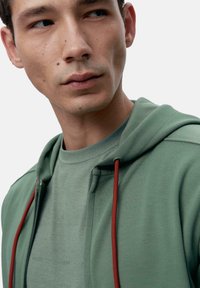 Hellgrüner Kapuzenpullover mit roten Kordeln, eng anliegendes Design, weiche Textur. Trägt einen passenden hellgrünen Shirt mit dezentem Branding.