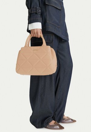Persona con completo in denim scuro che tiene una borsa trapuntata beige con manici superiori, indossando ballerine a punta con tonalità miste di marrone e viola.