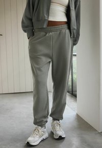 Pantaloni della tuta grigi chiari con vita elastica, tasche laterali e polsini, abbinati a scarpe da ginnastica bianche. Texture del tessuto morbida.