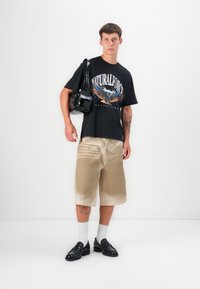 Sort grafisk t-shirt med ær design, beige pordoy-shorts, sorte loafers og hvide sokker. Modellen bærer en blank sort håndtaske.
