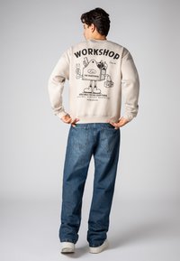 Cream sweatshirt featuring a graphic print on the back with text and cartoon character, paired with loose blue denim jeans and white sneakers.