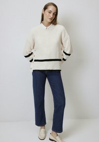 Pull crème en maille avec des rayures noires, détails boutons sur les côtés, associé à un jean taille haute bleu et des mocassins de couleur claire.