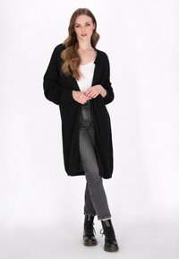 Langer schwarzer Cardigan aus strukturiertem Stoff, offen vorne, kombiniert mit einem weißen Oberteil und hoch geschnittenen grauen Jeans. Schwarze Schnürstiefel runden den Look ab.