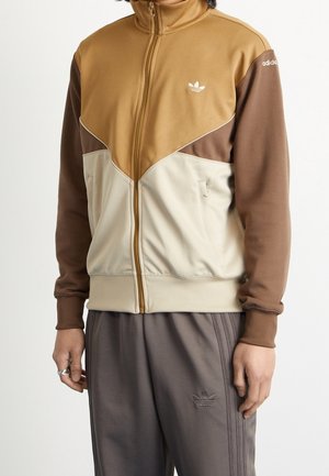 Person trägt eine braun-beige-tanfarbene Adidas Colorblock-Zipjacke mit passenden dunkelgrauen Adidas-Trainingshosen.