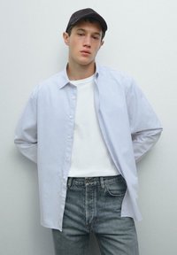 Overhemd met verticale lichtblauwe en witte strepen, gecombineerd met een effen wit T-shirt en grijze spijkerbroeken. Zwarte pet gedragen.