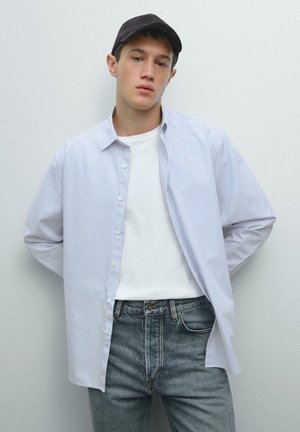 Camicia button-up con righe verticali azzurre e bianche, abbinata a una maglietta bianca e jeans in denim grigi. Cappellino nero indossato.