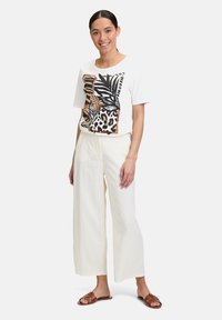 Weißes T-Shirt mit schwarzem und braunem Blatt- und Leopardenmuster, kombiniert mit cremefarbenen, weit geschnittenen Hosen und braunen flachen Sandalen.