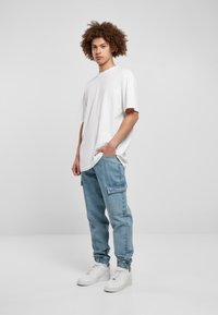 Hvid oversized t-shirt, lyseblå cargo-bukser med sidelommer, elastiske manchetter og hvide sneakers. Ensfarvet baggrund fremhæver fokus på tøjet.