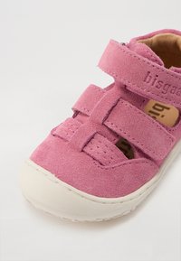 Roze nubuk sandala s dva podesiva Velcro remena, okruglim prstom i bijelim gumenim potplatom. Ime brenda ugravirano je na jednom remenu.