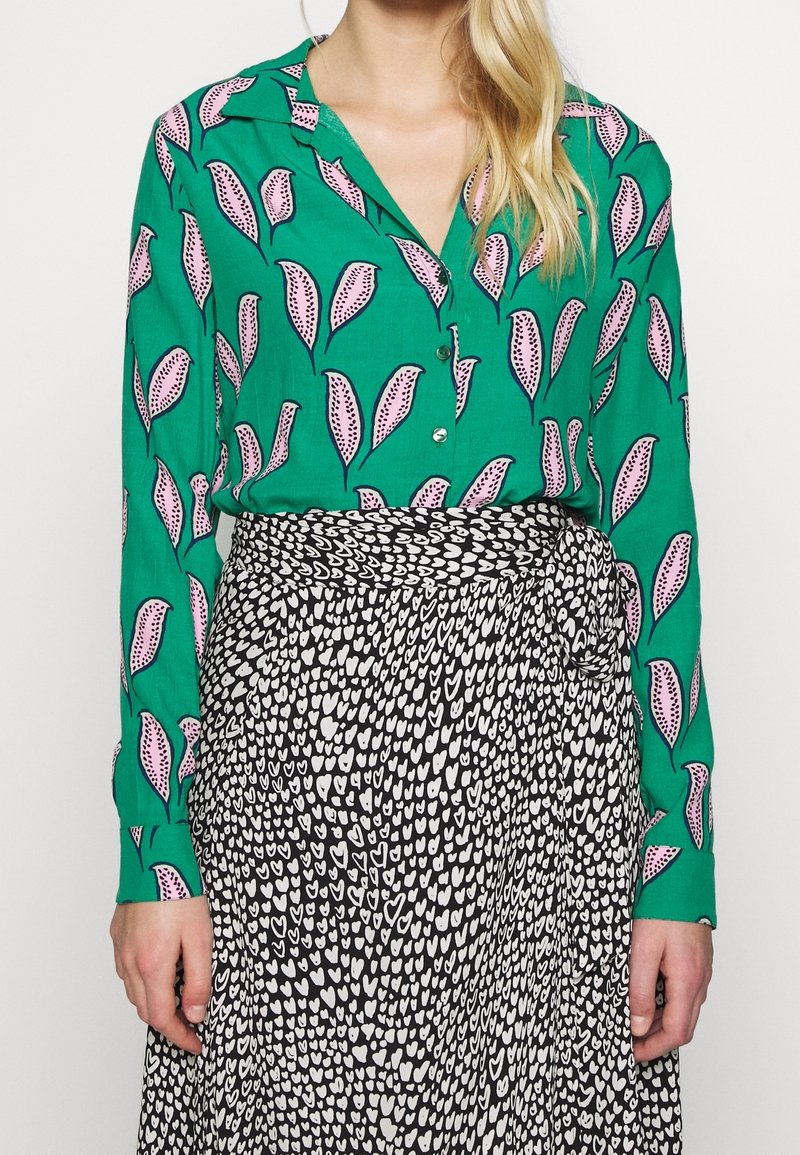 Blusa verde con motivo a foglie, con accenti rosa, abbinata a una gonna nera decorata con cuori bianchi; entrambi in tessuto morbido e fluido.