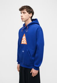 Νεαρός άνδρας που φοράει μπλε φούτερ με κουκούλα και πορτοκαλί τρίγωνο λογότυπο, καθώς και μαύρο παντελόνι, στέκεται μπροστά σε απλό λευκό φόντο.