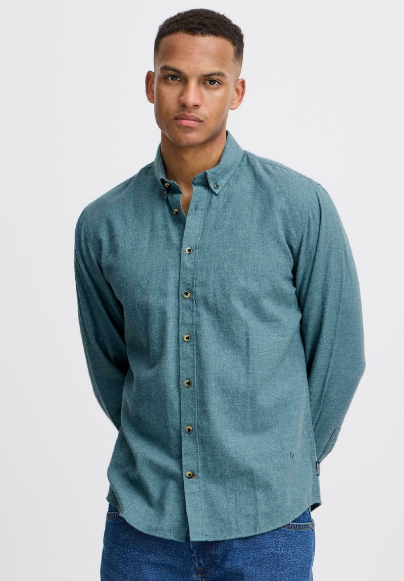 Chemise turquoise à boutons en tissu texturé avec manches longues et ourlet arrondi. Se distingue par des boutons dorés et un col pointu.