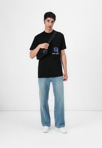 Black T-shirt with blue graphic print, paired with light blue straight-leg jeans and white sneakers. Crossbody bag in black material.