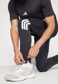 Melns sporta krekls ar mazu baltu logotipu, atbilstošas melnas bikses ar baltām svītrām un baltas sporta kurpes. Uzsvars uz piegriezumu un materiālu.