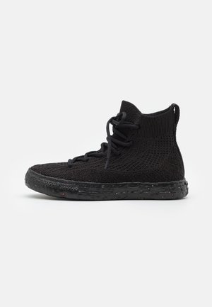 Baskets montantes noires en tissu tricoté, fermeture à lacets et semelle texturée avec de petites taches colorées, vues de côté sur fond blanc.