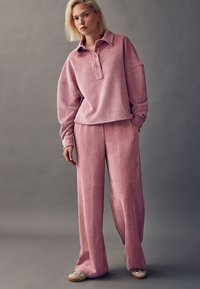 Pull-over en velours côtelé rose avec col et patte de boutonnage, associé à un pantalon ample assorti. Texture douce avec un motif côtelé subtil.