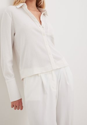 Kobieta ubrana w teksturowaną, kremowobiałą koszulę zapinaną na guziki, z szerokimi mankietami i luźne, białe spodnie z wysokim stanem na jednolitym tle.