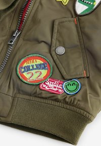 Veste bomber vert olive avec fermeture éclair à l'avant, poignets côtelés et divers patchs brodés aux motifs et textes colorés.