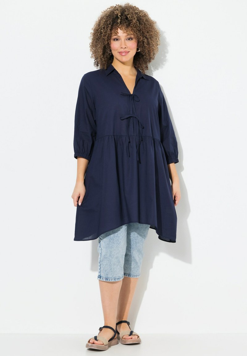 Marineblå bluse med løs pasform, skjortekrave, båndaccent og trekvartærmer, parret med lyse denim capri-bukser og sandaler.